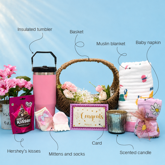 Baby shower gift basket set arranged on a light blue background with labeled callouts pointing to each item: insulated tumbler, basket, muslin blanket, baby napkin, flower pot with pink flowers, Hershey's kisses bag, mittens and socks, card, candle, and a pink packaged gift. No person, animal, or real environment is present, so this is Product/Packaging rather than Lifestyle.