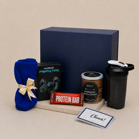 A curated gift set arranged on a beige surface with a large navy gift box behind it. The set includes a blue towel tied with a cream ribbon, a black HopUp Predator box, a red protein bar, a can of Brisket cranberry sauce cookies, a black shaker bottle, and a small card that says Cheers!.