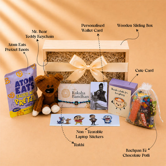 A curated gift box set arranged on a warm beige background with callout labels. The set includes a wooden sliding box with a cream ribbon, shredded paper filler, a personalized wallet card, a brown teddy bear keychain, a bag of Atom Eats Pretzel Knots, a cute card, a chocolate potli filled with colorful candies, and Rakhi stickers.