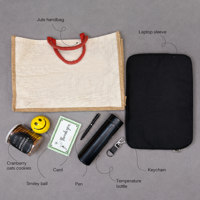 Collection of items including a jute handbag, laptop sleeve, cranberry oats cookies, smiley ball, card, pen, temperature bottle, and keychain on a gray background.