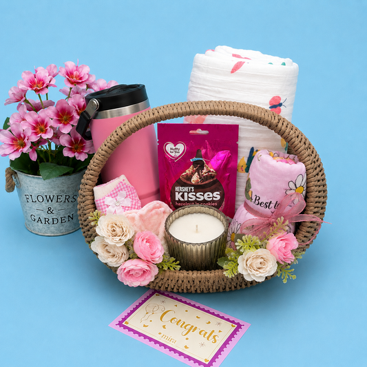 A baby shower gift basket arrangement on a light blue background, featuring a woven oval basket with pink and cream flowers, a pink candle, a pink travel tumbler, a bag of Hershey's Kisses, a folded pink cloth, and a stack of white diapers. A galvanized bucket planter with pink flowers sits to the left, and a congratulations card is placed in front.