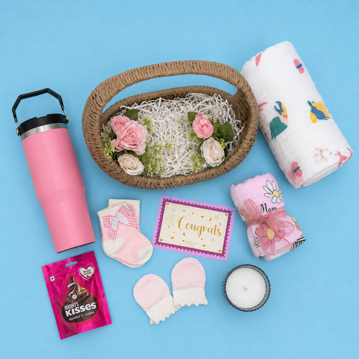 A baby gift set arranged on a light blue background, including a pink tumbler, a woven basket with flowers and shredded paper, a white muslin swaddle with colorful prints, a pink and white bootie, a pink and white card, a pink flower-wrapped item, a small candle, and a pink chocolate kiss packet.