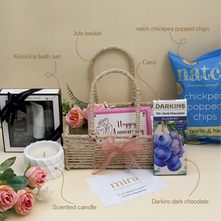 Gift basket with Kimirica bath set, Jute basket, Darkins dark chocolate, natch chickpea popped chips, and scented candle on a beige surface.