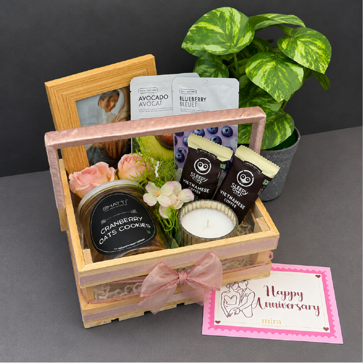 Gift basket with candles, snacks, and a photo frame on a gray surface with a plant in the background.