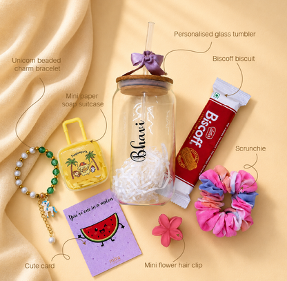 A styled flat-lay of a personalized glass tumbler with a wooden lid and purple bow, surrounded by labeled gift items including a unicorn beaded charm bracelet, a mini paper suitcase, a Biscoff biscuit, a scrunchie, a watermelon card, and a mini flower hair clip on a beige satin background.