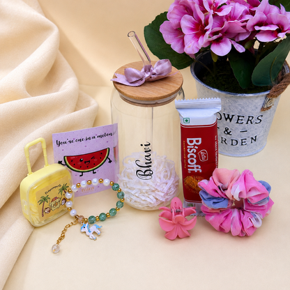 A styled flat-lay of a gift set on a beige fabric background, featuring a clear glass jar with a bamboo lid and purple bow, a tube of Biscoff chocolates, a small yellow suitcase, a beaded bracelet, and pink flowers.