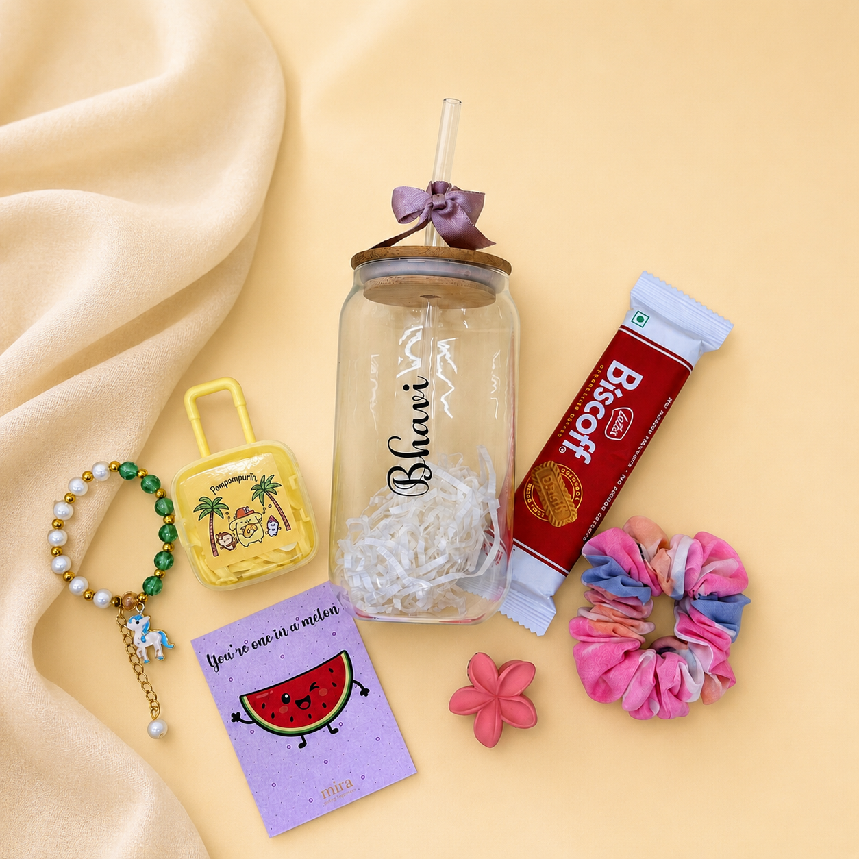 A styled flat-lay of a clear glass jar tumbler with a bamboo lid and straw, decorated with a purple bow and the word 'Bhari' printed on it. The tumbler is surrounded by various colorful accessories, including a pink and blue scrunchie, a green beaded bracelet, a yellow cartoon pouch, a purple card with a watermelon illustration, a red Biscuit tube, and a small pink flower, all arranged on a beige satin fabric background.