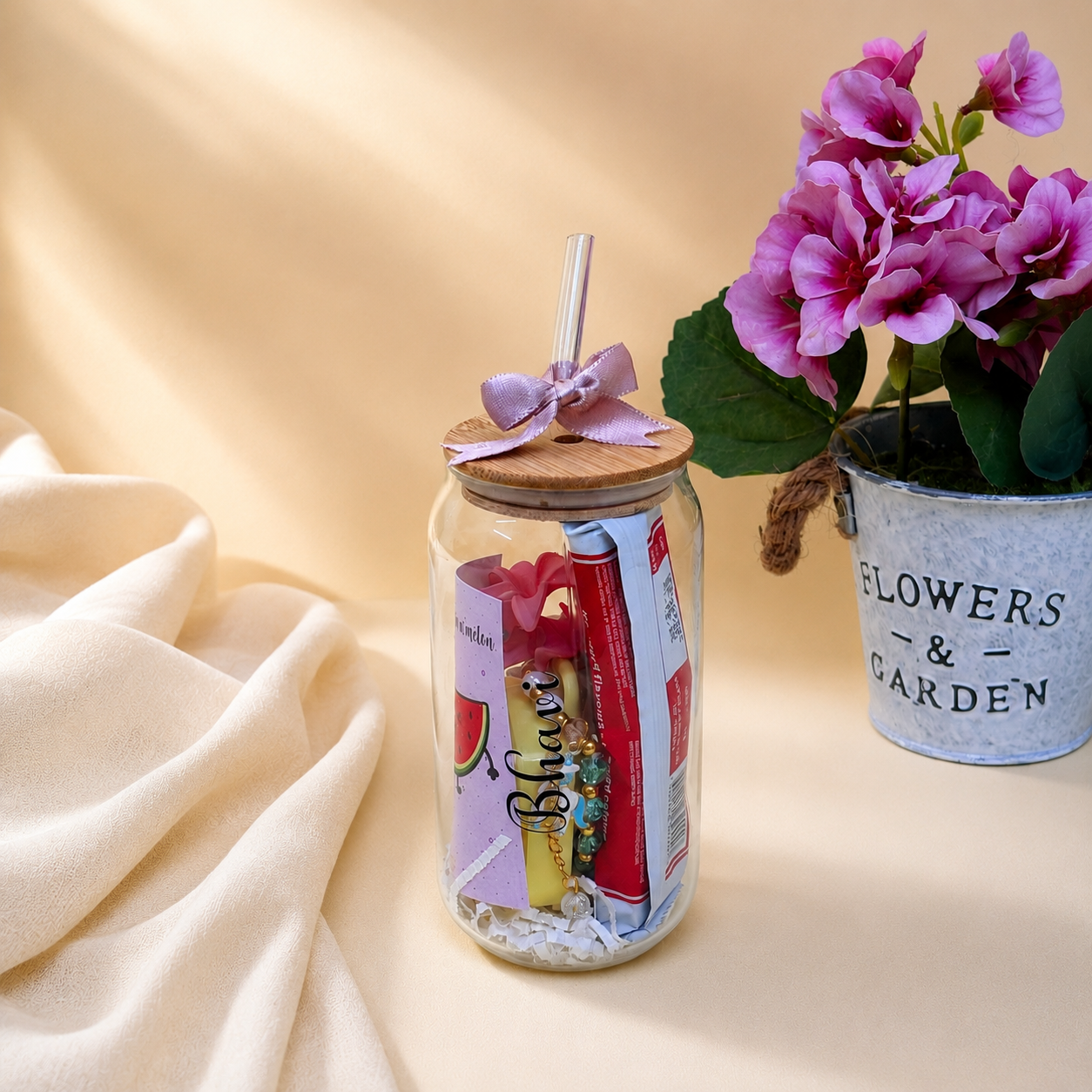 A clear glass jar gift container with a wooden lid and a lavender ribbon bow, filled with assorted packaged snacks and accessories. The jar sits on a draped cream fabric surface next to a potted plant of pink flowers in a metal bucket labeled 'FLOWERS & GARDEN'.
