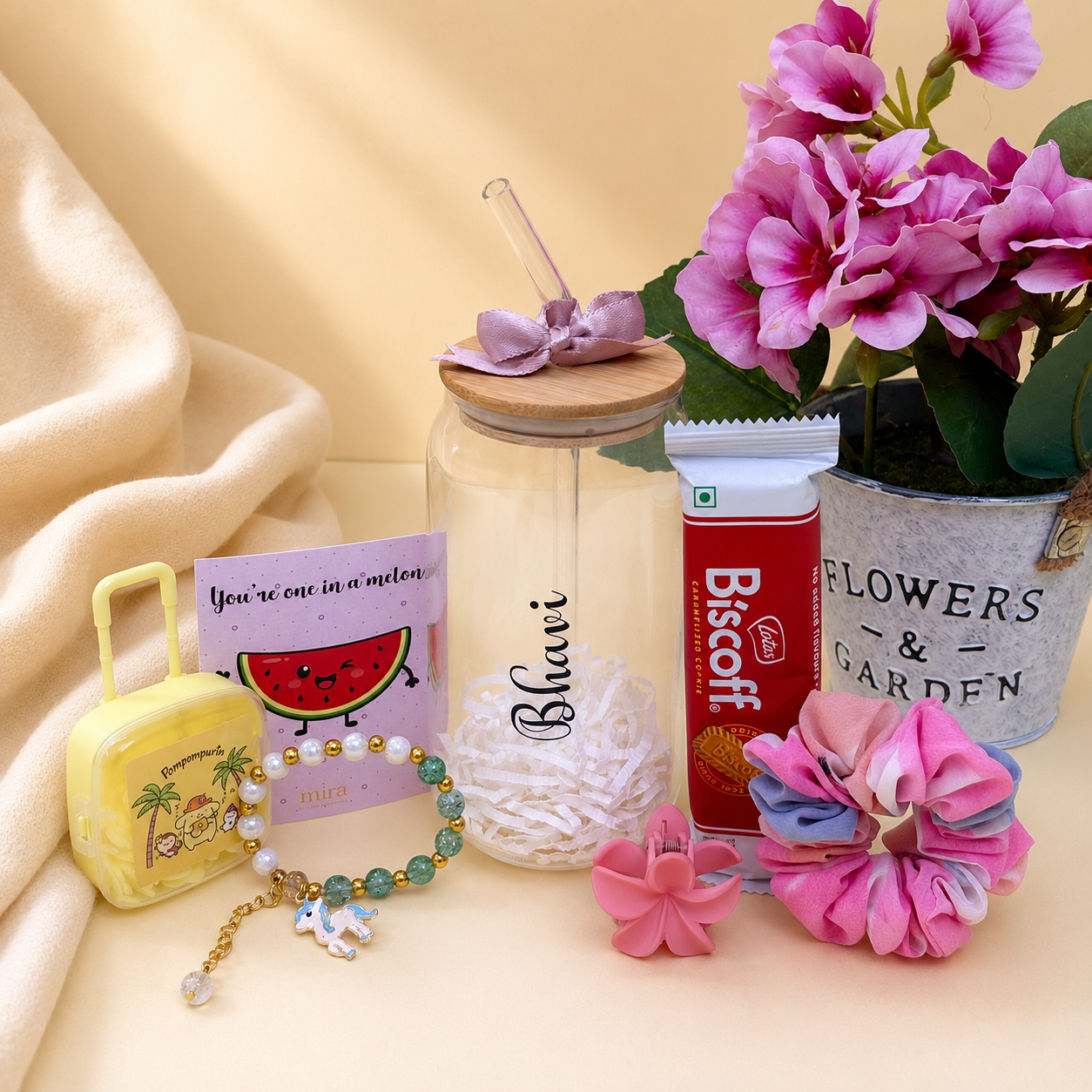 A styled flat-lay of a gift set on a warm beige background, featuring a clear glass bottle with a wooden lid and purple bow, a yellow cartoon suitcase charm, a beaded bracelet, a watermelon card, a Biscotti tube, and pink flowers.