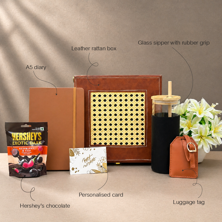 Collection of travel-related items including a leather rattan box, glass sipper with rubber grip, A5 diary, Hershey's chocolate, personalized card, and luggage tag on a neutral background.