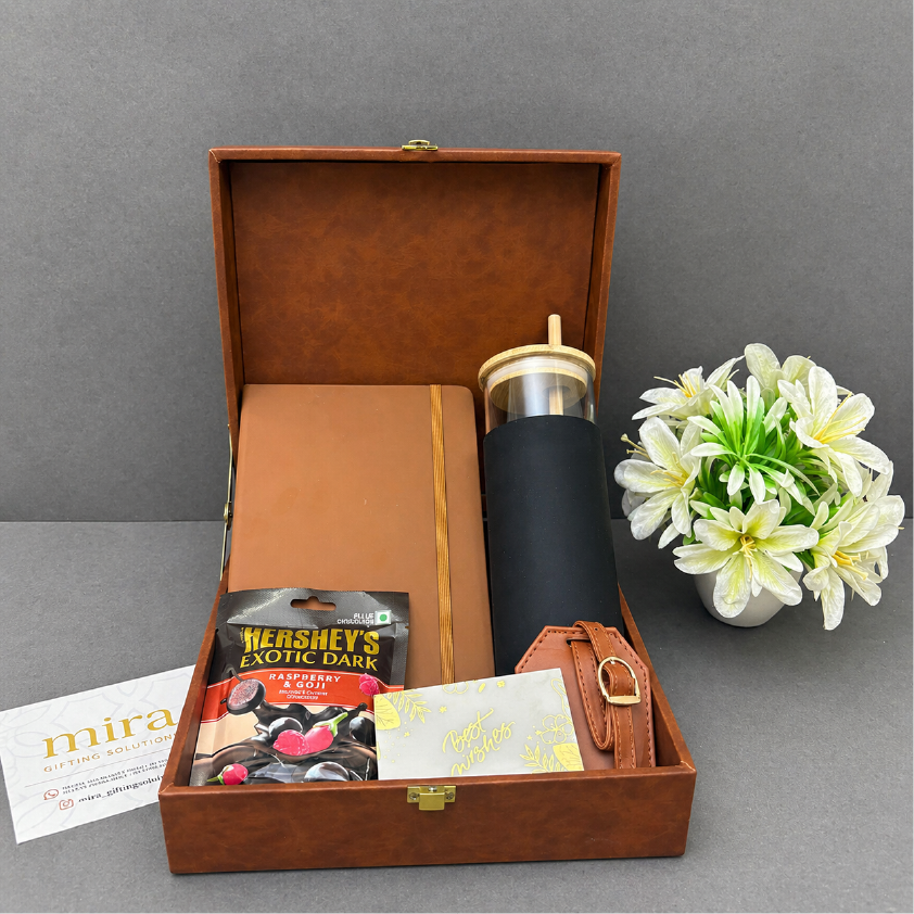Gift set with a black tumbler, notebook, and chocolate bars in a brown box on a gray surface.