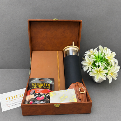 Gift set with a black tumbler, notebook, and chocolate bars in a brown box on a gray surface.