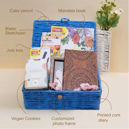 Gift basket with stationery items including a mandala book, water sketchpen, vegan cookies, and a printed cork diary on a beige background.