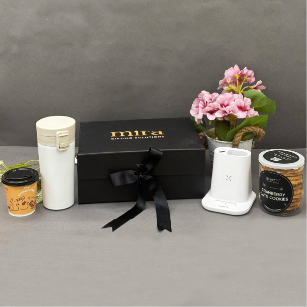 Gift set with a black box, white mug, coffee cup, small plant, and cookies on a gray surface.