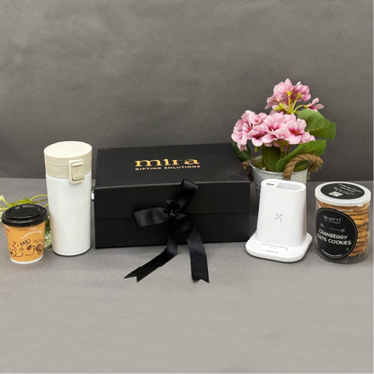Gift set with a black box, white mug, coffee cup, small plant, and cookies on a gray surface.