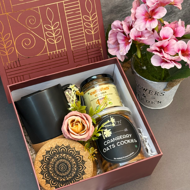 Gift box with a black mug, cookies, and a plant on a gray surface