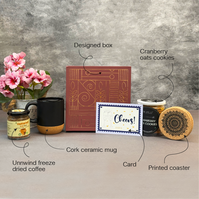 Gourmet gift set with a designed box, cork ceramic mug, printed coaster, card, cranberry oats cookies, and unwind freeze-dried coffee on a gray surface.