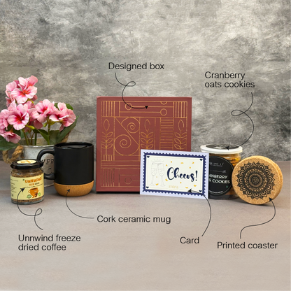 Gourmet gift set with a designed box, cork ceramic mug, printed coaster, card, cranberry oats cookies, and unwind freeze-dried coffee on a gray surface.