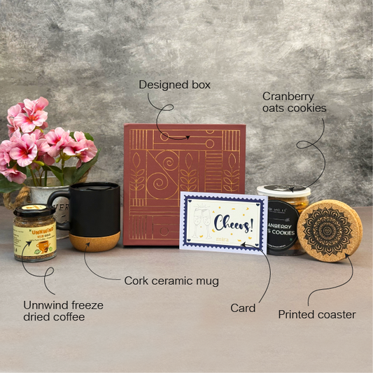Gourmet gift set with a designed box, cork ceramic mug, printed coaster, card, cranberry oats cookies, and unwind freeze-dried coffee on a gray surface.