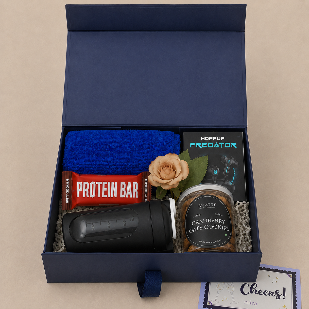 A curated gift box set displayed in an open navy blue box on a beige background. The set includes a blue towel, a red protein bar, a black shaker bottle, a tin of cranberry oats cookies, a black Hoppup Predator tumbler, and a peach-colored rose.