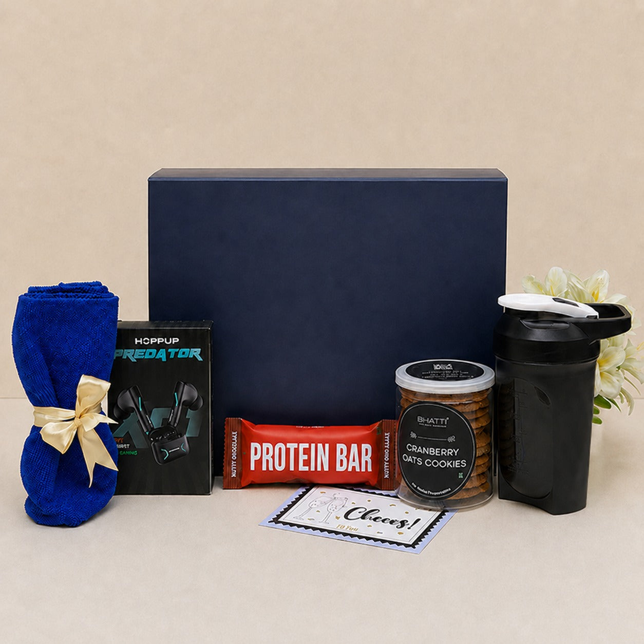 A gift set arranged in front of a large navy blue box on a beige background. The set includes a rolled blue towel tied with a cream ribbon, a black HOPUP Predator tube, a red protein bar, a jar of cranberry ginseng cookies, a black shaker bottle, and a small card that says 'Cheers!'.