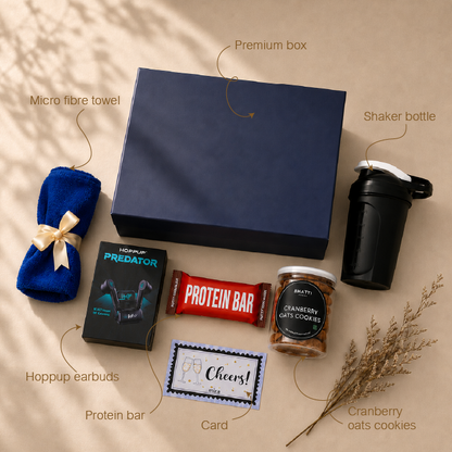 A curated gift box set arranged on a beige surface with handwritten callouts naming each item: a navy premium box, a blue microfiber towel tied with a cream ribbon, a black shaker bottle, a black Hoppup Predator earbud box, a red protein bar, a jar of cranberry oats cookies, and a small card. Decorative dried stems sit at the lower right.