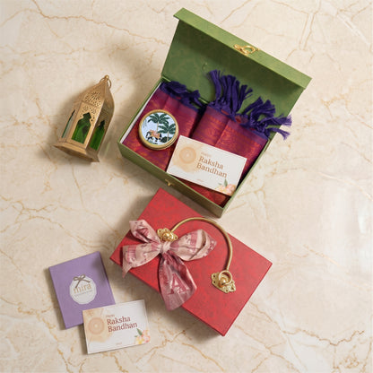 A gift set arranged on a light beige marble surface, featuring an open green box with purple tassels and a small round decorative item, a red gift box tied with a pink patterned ribbon and gold clasp, a small lantern, and two branded cards reading Raksha Bandhan.