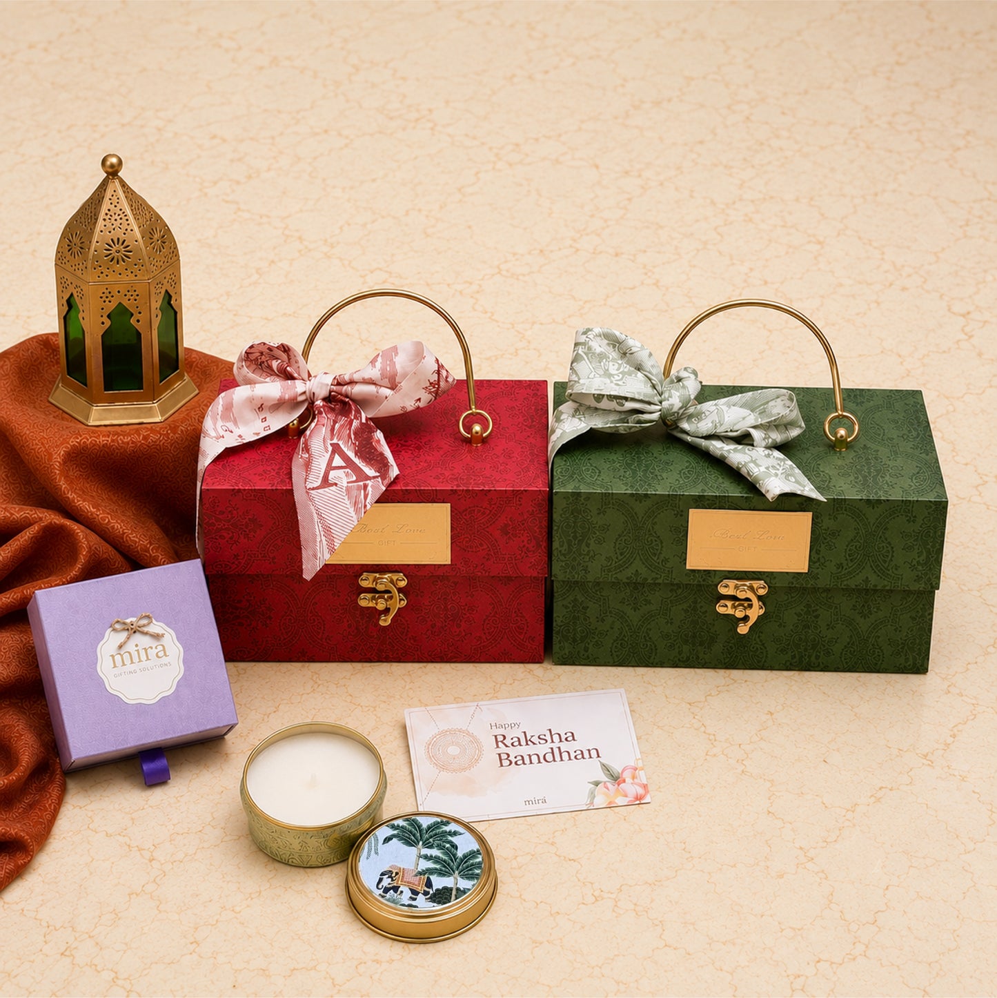 A styled gift set on a beige textured background featuring two ornate rectangular gift boxes with gold handles and clasp lids, one red and one green, both tied with decorative satin bows. A purple box labeled 'mima', a round tin candle, and a small card reading 'Rakesh Bandhan' are arranged in front.