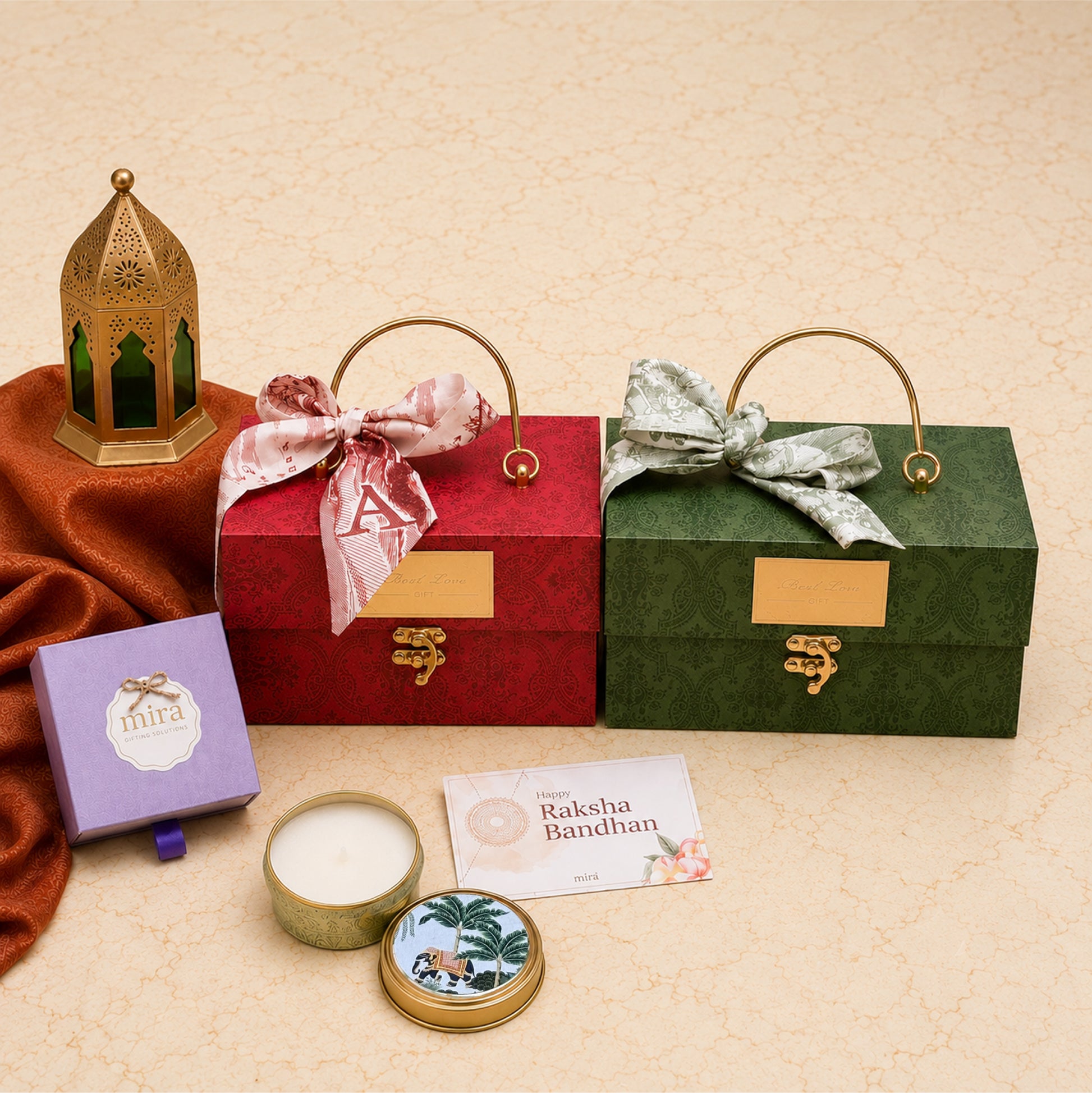 A styled gift set on a beige textured background featuring two ornate rectangular gift boxes with gold handles and clasp lids, one red and one green, both tied with decorative satin bows. A purple box labeled 'mima', a round tin candle, and a small card reading 'Rakesh Bandhan' are arranged in front.