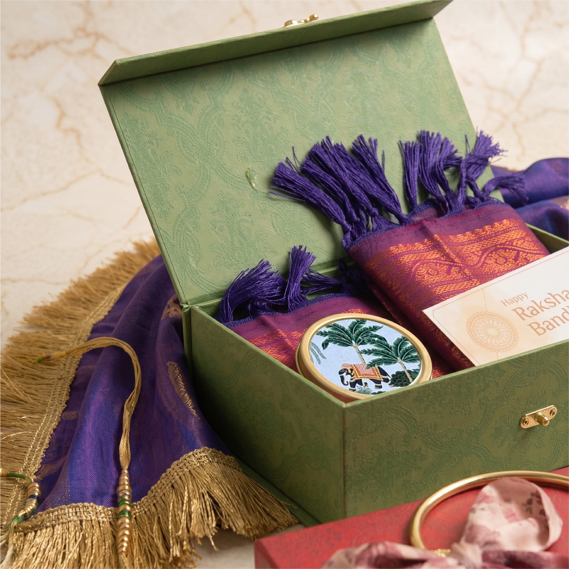 A green gift box is open to reveal a purple and gold Raksha Bandhan scarf with tassels, a small round decorative tin, and a card reading 'Happy Raksha Bandhan.' A matching purple scarf with gold fringe is draped beside the box on a light marble surface.
