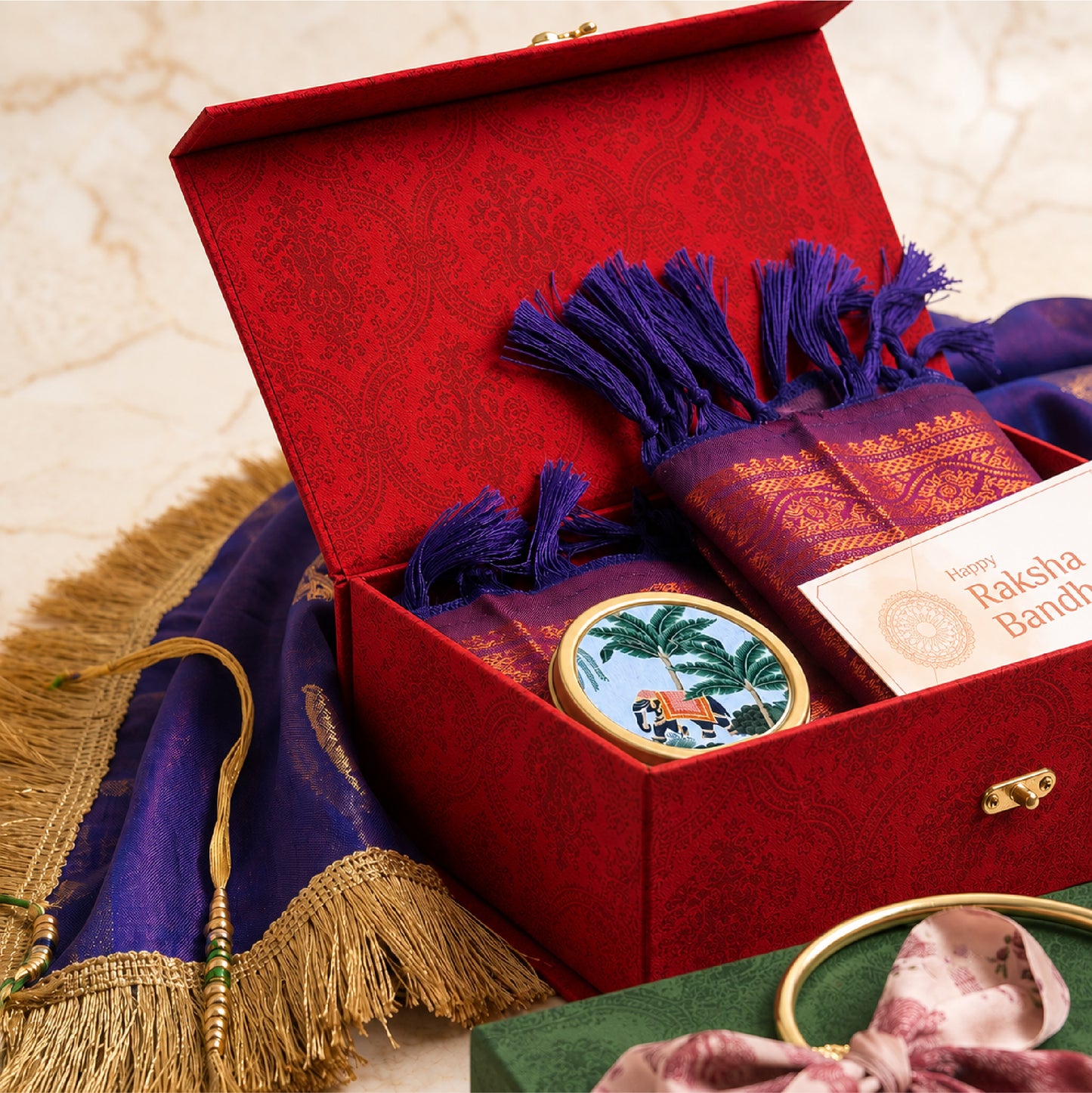 A red ornate gift box opened to reveal a purple and gold shawl, a small round decorative item with a palm tree illustration, and a card that reads 'Happy Raksha Bandhan.'.