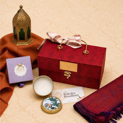 A Raksha Bandhan gift set arranged on a beige patterned surface, featuring a red patterned gift box with a gold clasp and pink satin bow, a purple Mirza candle box, a small round tin, a folded red shawl, and an orange fabric cloth.