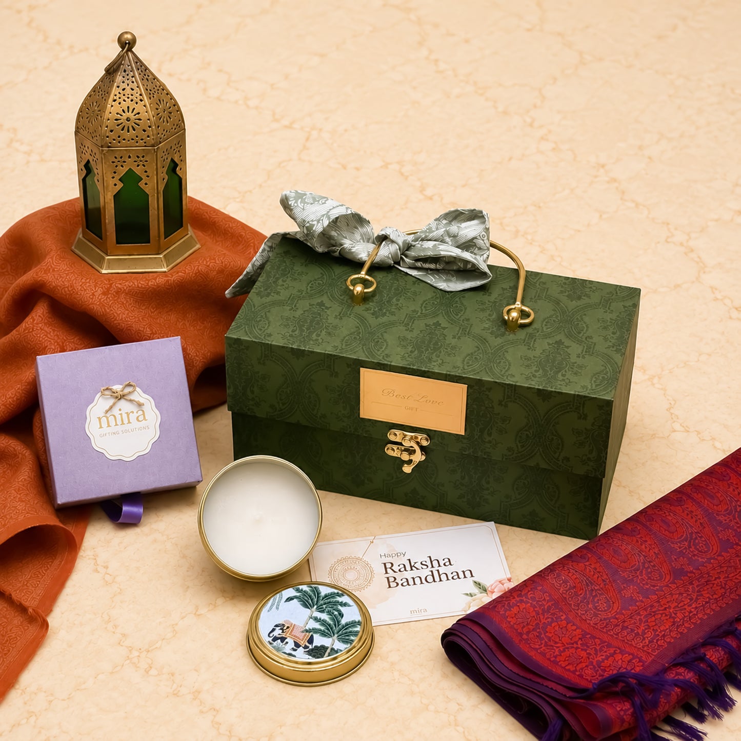 A curated gift set arranged on a beige textured surface, featuring a dark green patterned presentation box with gold-tone handles and clasp, a silver bow, a small lavender box labeled 'mira', an open round tin with white wax, a card reading 'Raksha Bandhan', a red patterned scarf, and an ornate gold lantern with green glass panels.