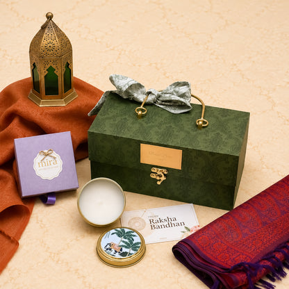 A curated gift set arranged on a beige textured surface, featuring a dark green patterned presentation box with gold-tone handles and clasp, a silver bow, a small lavender box labeled 'mira', an open round tin with white wax, a card reading 'Raksha Bandhan', a red patterned scarf, and an ornate gold lantern with green glass panels.