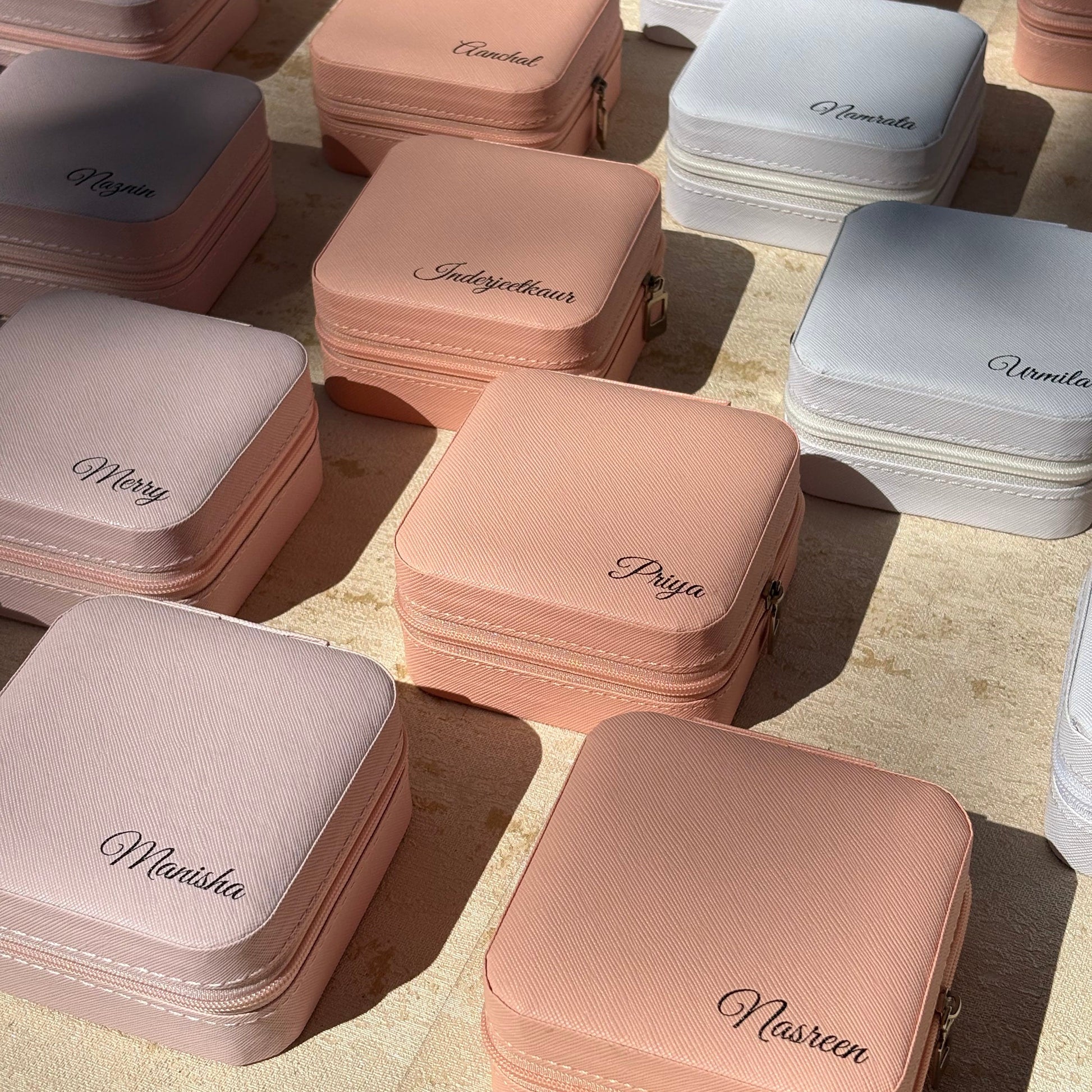 Multiple small zippered cosmetic or jewelry cases arranged on a light beige surface, shown in soft blush pink, white, and pale gray. Each case has a script brand name printed on the front.