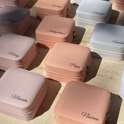 Multiple small zippered cosmetic or jewelry cases arranged on a light beige surface, shown in soft blush pink, white, and pale gray. Each case has a script brand name printed on the front.