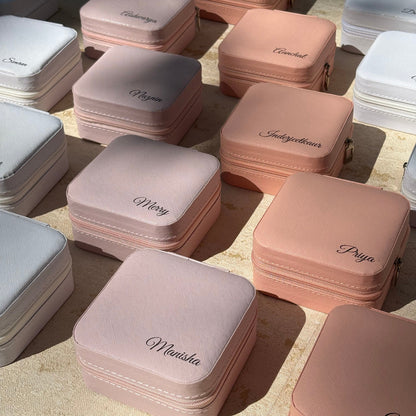 A collection of small, square, zippered jewelry boxes in soft pastel shades of pink, peach, and white, arranged on a light beige stone surface. Each box is personalized with an elegant black cursive name, such as 'Manisha', 'Priya', and 'IndigoBour'.