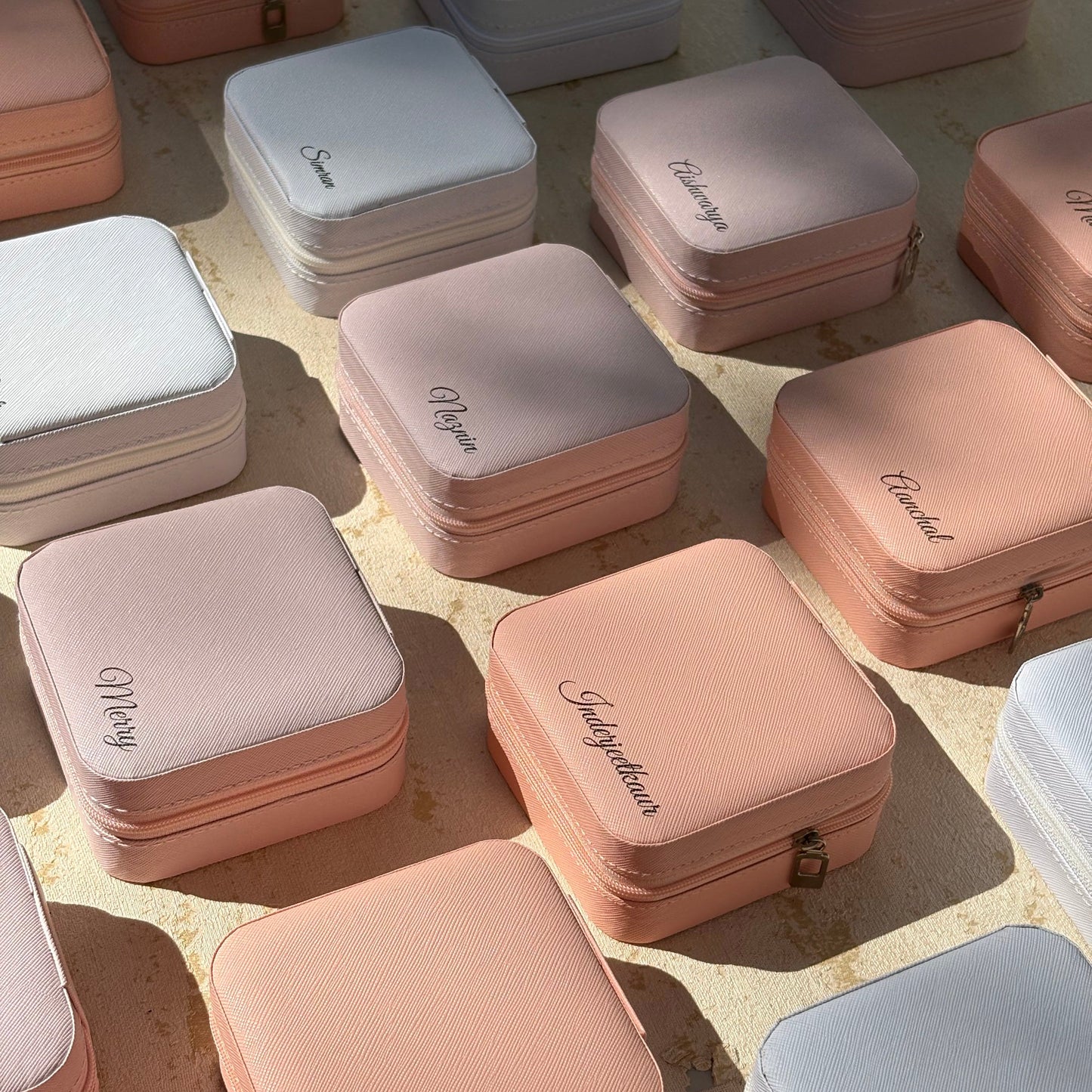 A collection of small square zippered jewelry boxes in pastel shades of pink, peach, white, and light blue, arranged on a textured beige surface with natural sunlight casting shadows.