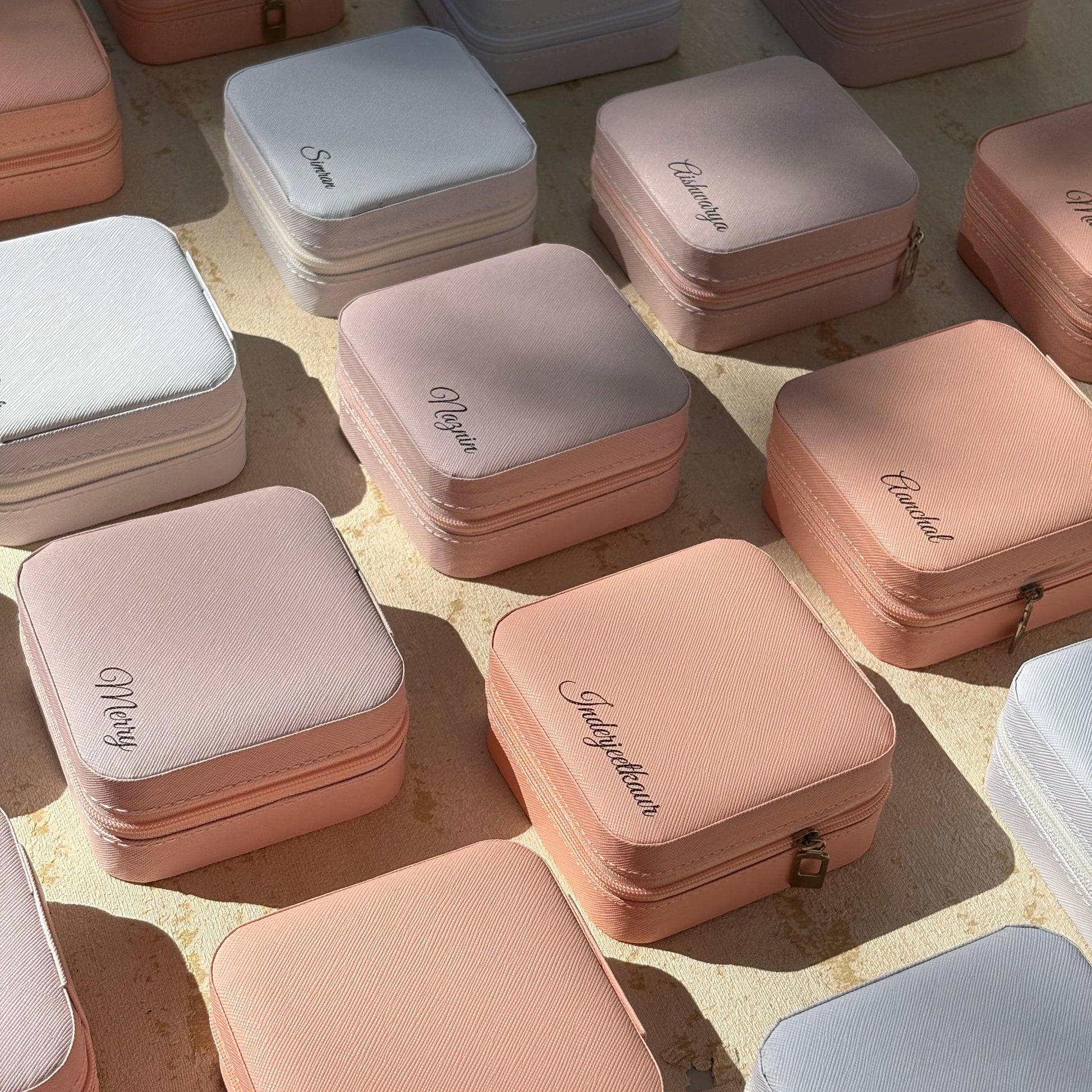A collection of small square zippered jewelry boxes in pastel shades of pink, peach, white, and light blue, arranged on a textured beige surface with natural sunlight casting shadows.