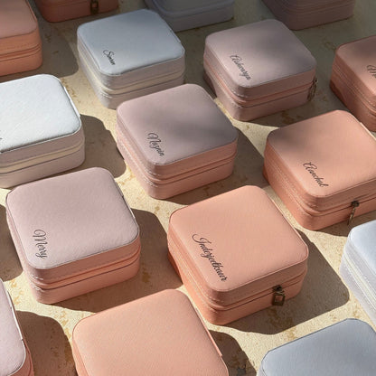 A collection of small square zippered jewelry boxes in pastel shades of pink, peach, white, and light blue, arranged on a textured beige surface with natural sunlight casting shadows.