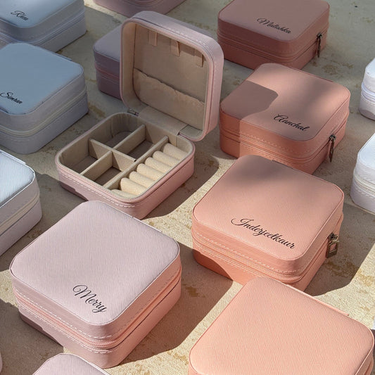 A collection of small square jewelry boxes in soft pastel shades of pink, peach, lavender, and white, arranged on a sunlit stone surface. Several boxes are open, showing interior compartments and ring slots, while others are closed with embroidered names such as Mary, Nicole, and Priya visible on the front.