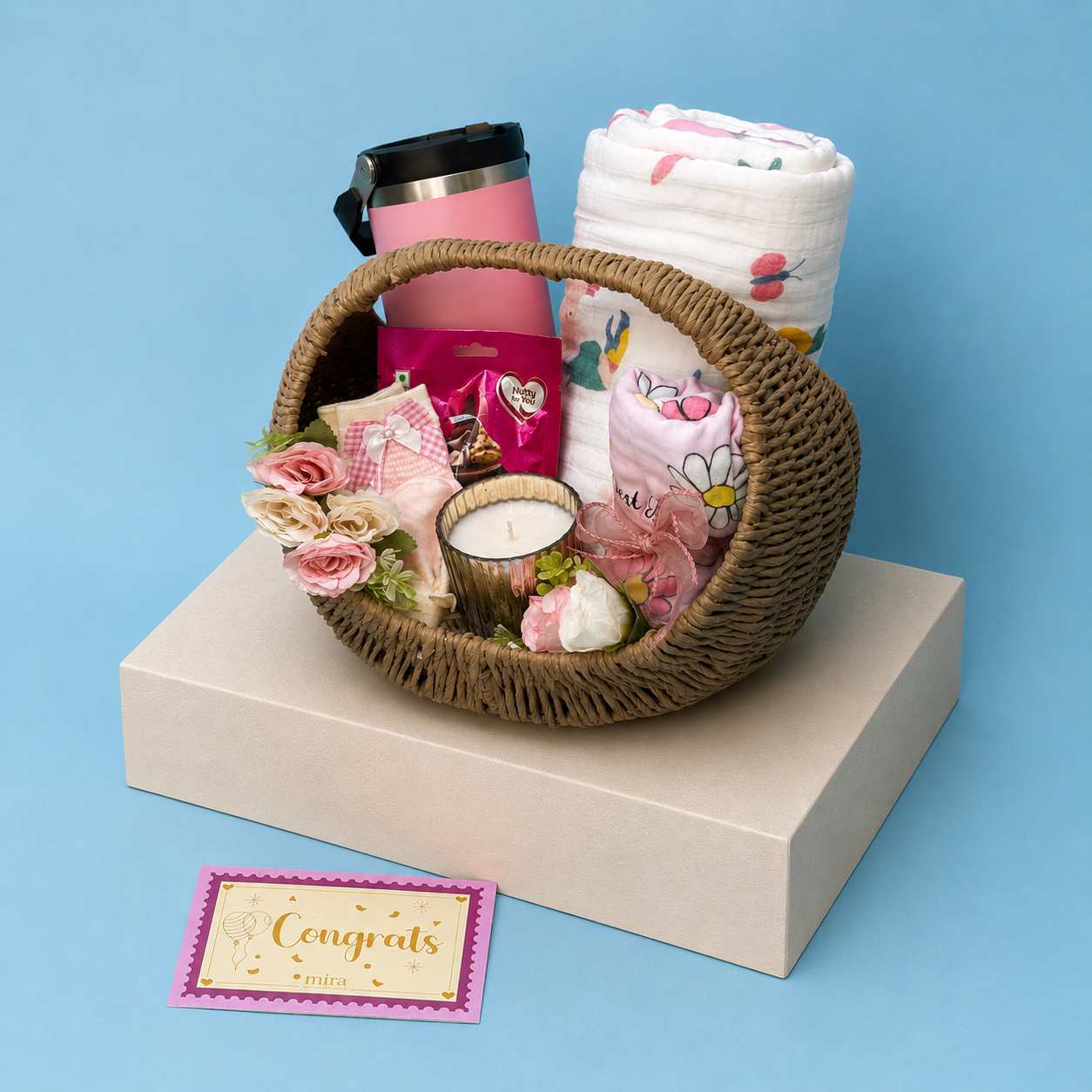 A baby shower gift basket arranged on a cream-colored box against a solid blue background. The basket contains a pink travel mug, a white and pink swaddle blanket, a pink and white baby bib, a small pink and white plush toy, a candle, and pink roses.