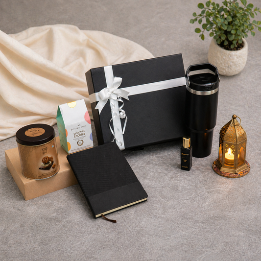 Gifting set with black box, notebook, travel mug, and candles on a gray surface.