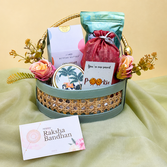 A Raksha Bandhan gift basket set on draped sage green fabric, featuring a mint green basket with a gold handle, filled with assorted treats including butter sweets, a cookie, and a pink drawstring pouch, decorated with roses and gold-toned floral accents. A Raksha Bandhan greeting card is placed in front.