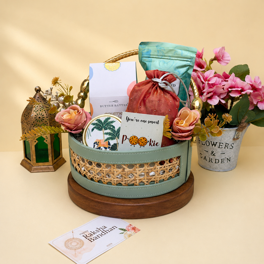 A curated gift basket arrangement on a wooden base, featuring a mint green basket with a woven gold pattern, a pink pouch, a white box labeled Butter Batty, a round palm-themed coaster, a small card reading 'You're too precious', and decorative flowers and lanterns around it on a beige studio background.
