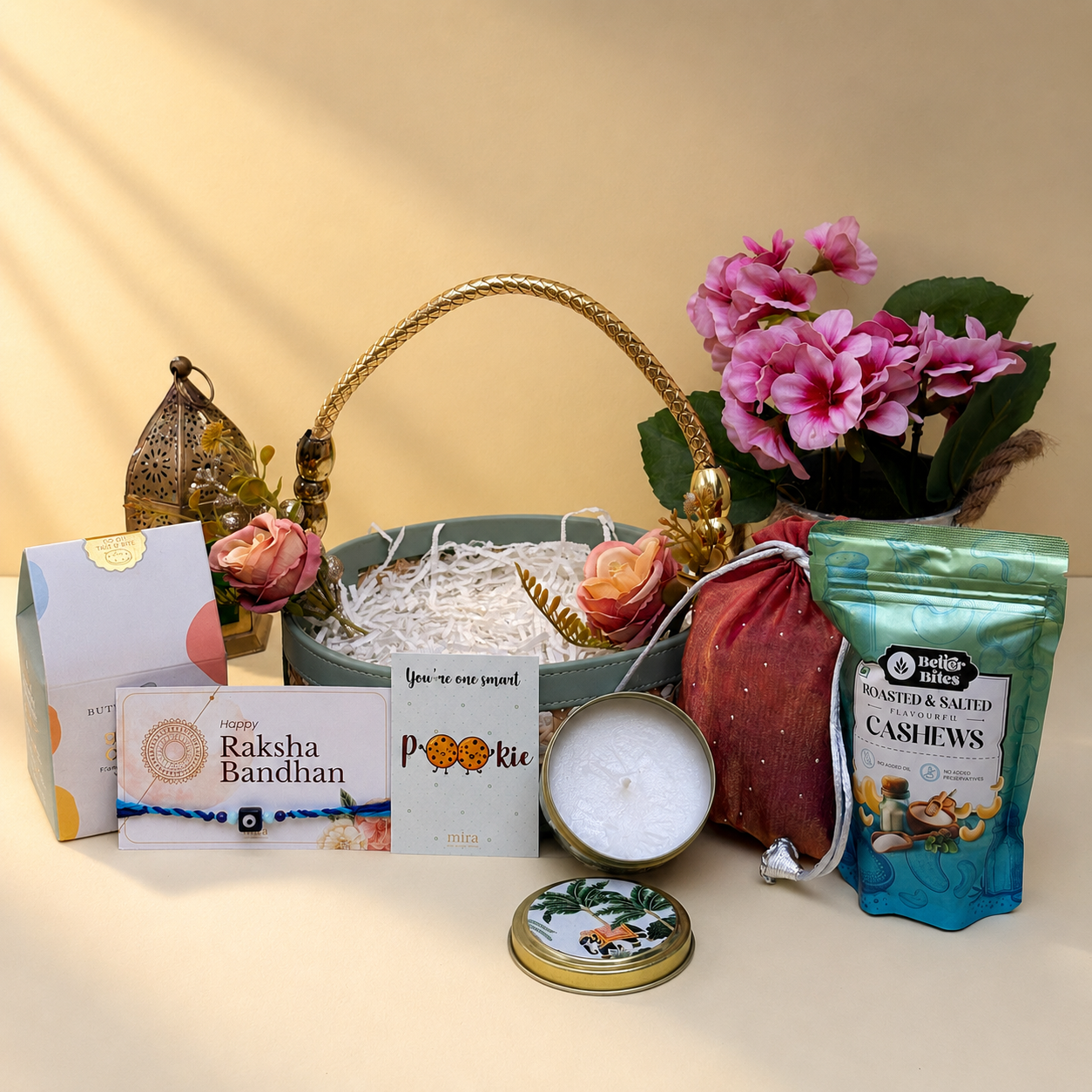 A styled gift basket arrangement on a beige background, featuring a mint green basket with a gold handle, pink flowers, a gold decorative lantern, a white gift box, a card labeled Raksha Bandhan, a small candle, a red drawstring bag, and a blue pouch of Cashews by Believable.