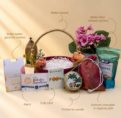 A curated gift basket arrangement on a warm beige background with handwritten callout labels pointing to each item. The set includes a red basket with a gold handle, pink flowers, a green packaged cashew product, a tin candle, a pink drawstring pouch, a card, a butter batter cookie box, and a decorative box.
