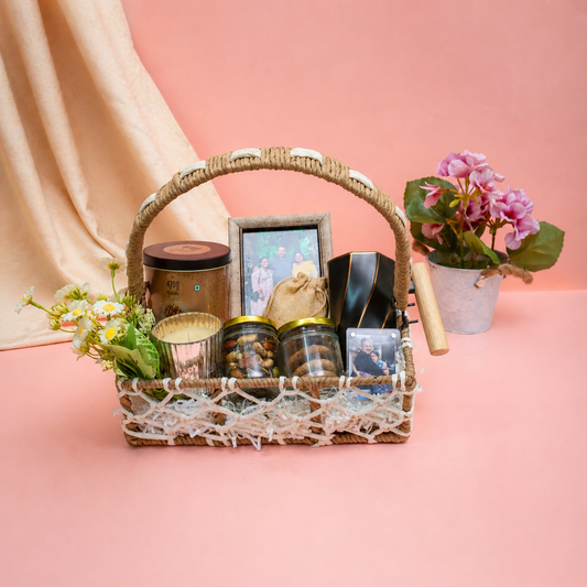 Wicker gift basket with various items including jars, a photo frame, and a plant on a pink background