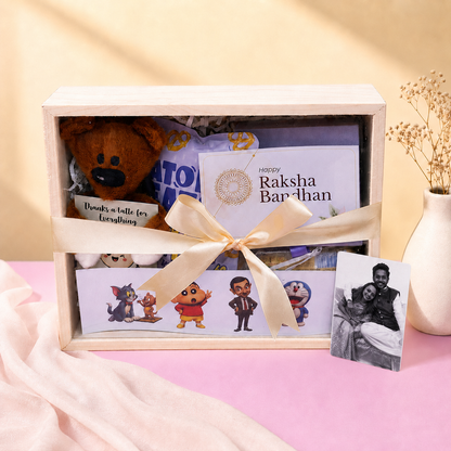 A wooden gift box tied with a cream ribbon, filled with assorted surprises including a brown plush bear, a greeting card reading 'happy Raksha Bandhan,' character strips, and a small black-and-white photo card, styled on a pink surface with a beige background and dried flowers in a vase.