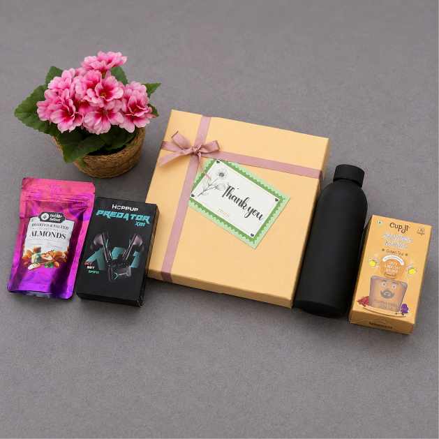 Gift box with pink ribbon, small plant, snacks, and a bottle on a gray surface